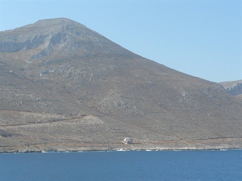 Arid rocky mountain above blue sea, a small white house on the shore