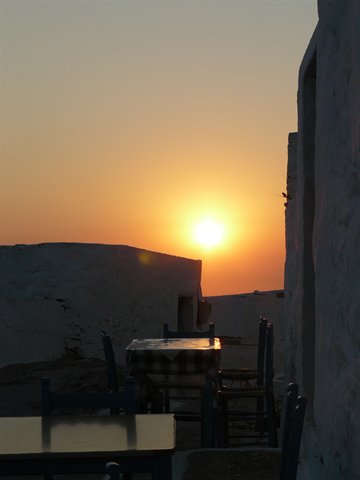 Wooden tables and chairs among whitewashed walls, orange sun setting over the sea