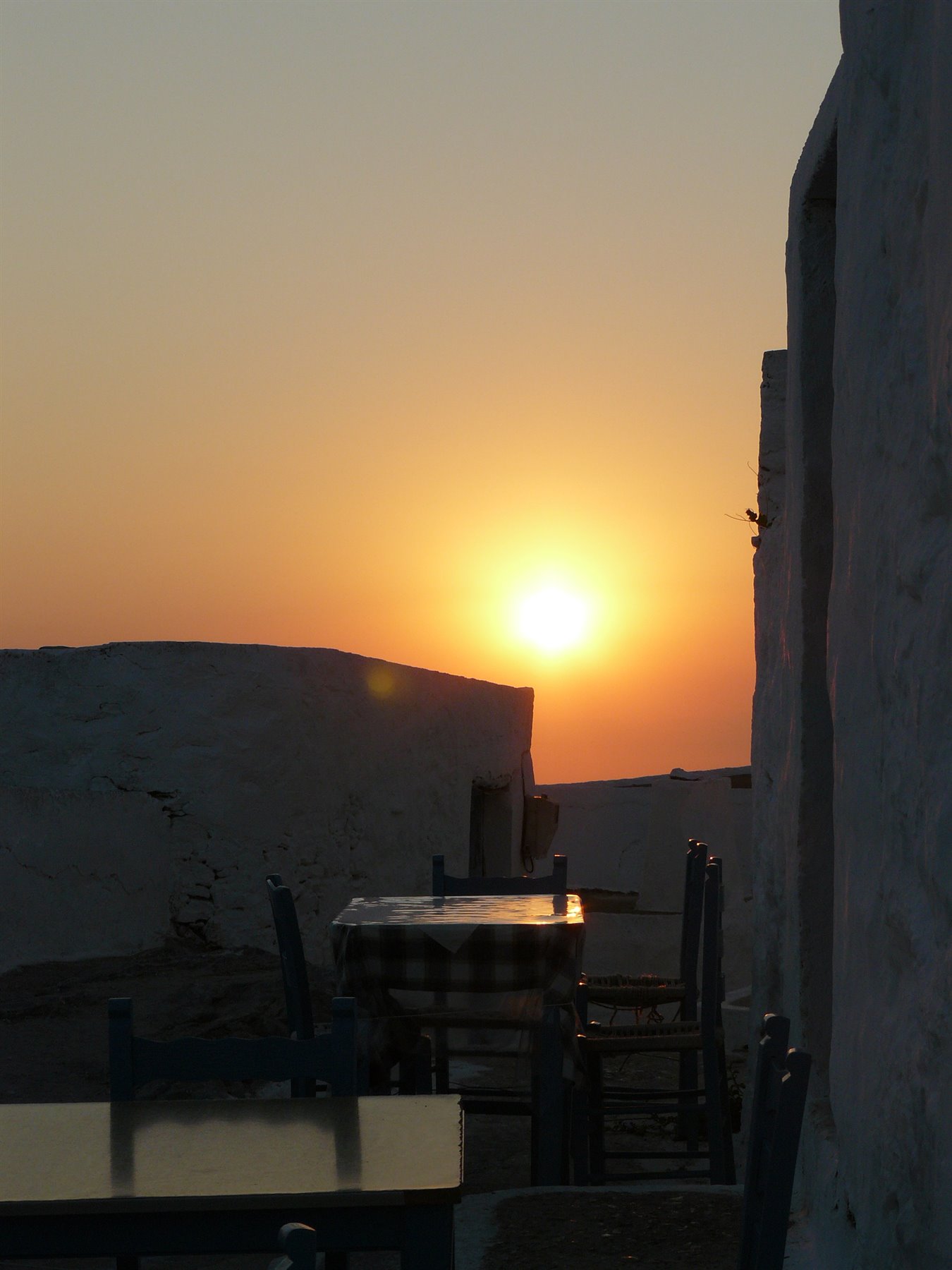 Tavoli e sedie di legno tra muri bianchi a calce, il sole arancio scende