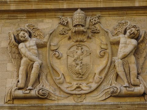Two carved stone angels flank a papal coat of arms on a facade