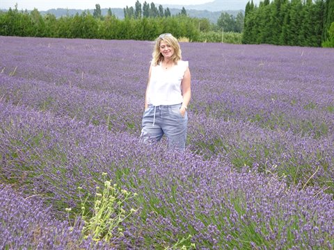 Blonde woman in white top standing among blooming lavender rows, trees behind
