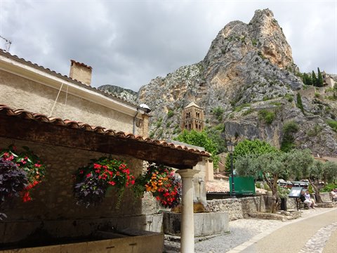 Hanging flowers over stone fountain, church tower and cliff in Moustiers-Sainte-Marie