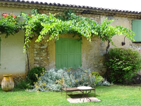 Vine over stone house with green shutters, wrought-iron bench on lawn