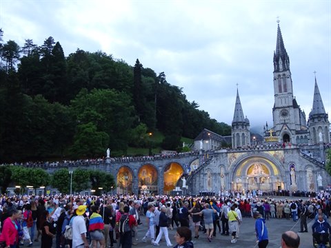 Pilgrim crowd before the Lourdes basilica, candles glowing at dusk