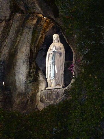 White statue of the Virgin in the Lourdes grotto, lit among rock and ivy