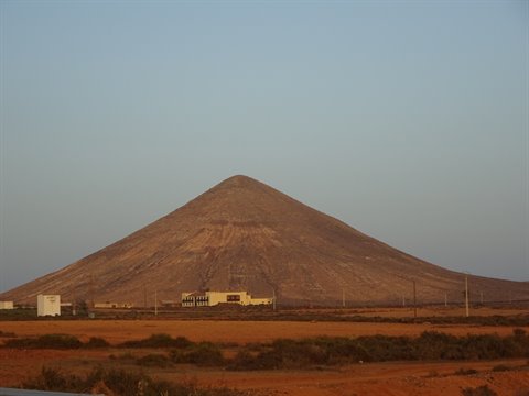 Dry volcanic cone rising over reddish fields and small buildings, pale evening sky