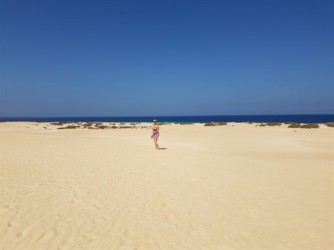 Woman walks on the pale sand dunes of Corralejo, blue sky and ocean behind