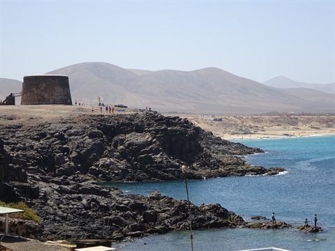 Old stone tower on volcanic coast at El Cotillo, tourists among black rocks