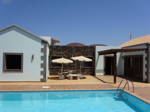 Blue pool in front of whitewashed house with lava stone wall, umbrellas and wooden pergola