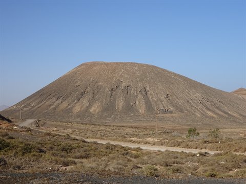 Arid volcanic cone above a dirt track with sparse shrubs under blue sky
