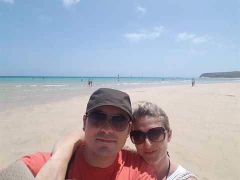 Couple selfie in cap and sunglasses on sandy beach with kitesurfers in the distance