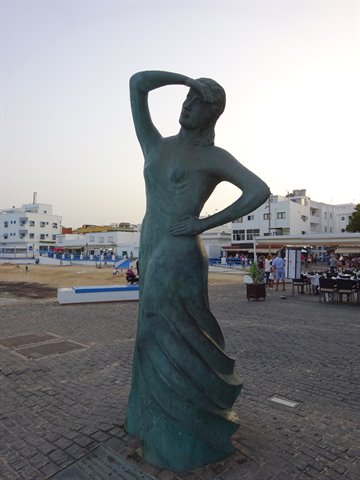 Green bronze statue of a woman on a plaza by the beach, whitewashed buildings