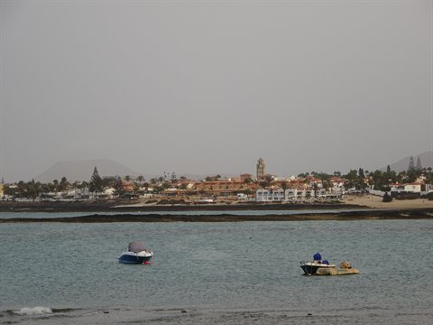 Two motorboats on turquoise water in front of white houses and a tower, Corralejo