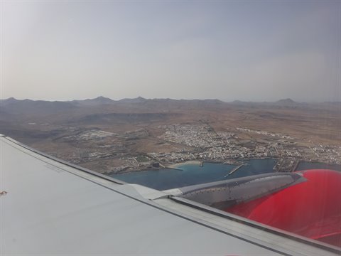 From the plane, the red wing flies over a coastal town among arid volcanic hills