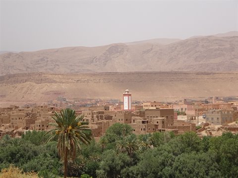 Minareto bianco sopra le case di terra, palme e montagne aride a Tinghir