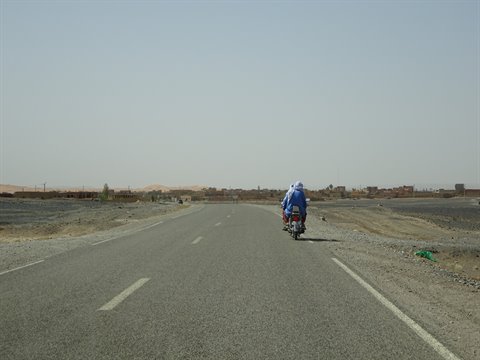 Blue-clad motorcyclist on paved road heading toward Merzouga dunes