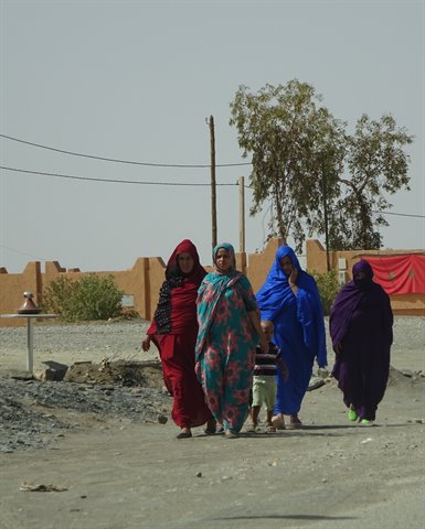 Veiled women in colorful kaftans walk with a child on sandy road