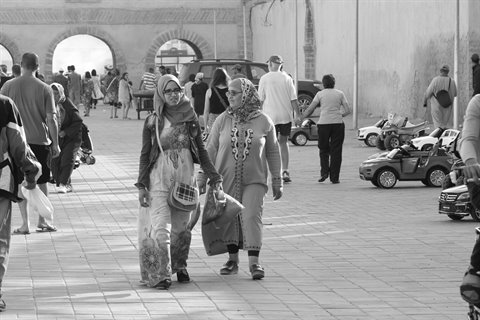 Two women walk under the arches of a busy square in black and white