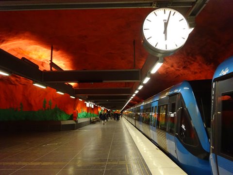 Blue train stopped at Solna Centrum station, red rocky ceiling, large clock