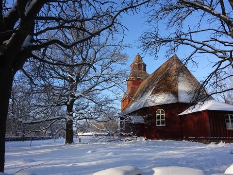 Snow-covered red wooden church at Skansen, bare branches against blue sky