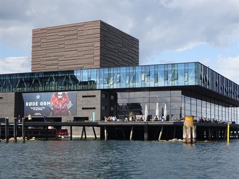 Copenhagen Royal Playhouse on stilts, glass and rust-brown brick, people at waterfront terrace