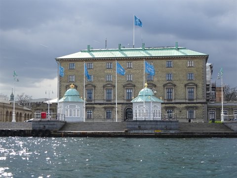 Waterfront palace with green domed pavilions, blue flags, cloudy sky