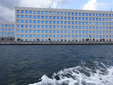Grey building with blue windows along the quay, people walking, white wake on water