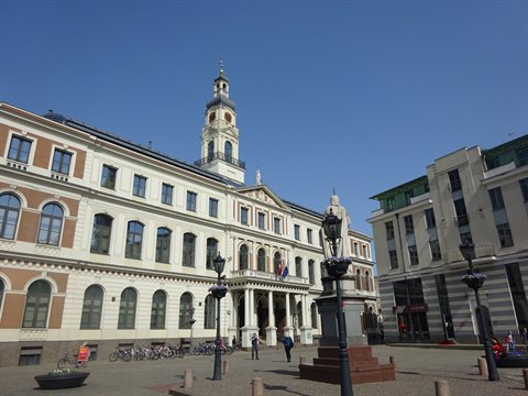 Riga town hall square, white building with steeple, statue and bicycles under sunny sky