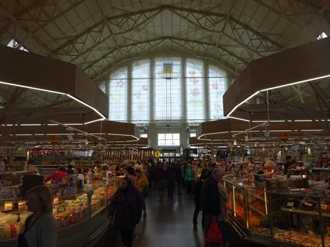 Indoor market with iron trusses and stained-glass window, shoppers browsing food stalls