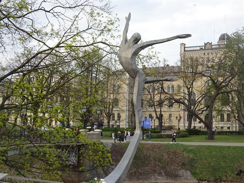 Metal sculpture of a leaping dancer on a bridge, trees and historic building