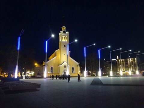 Illuminated yellow church at night in a square with blue lampposts, people walking