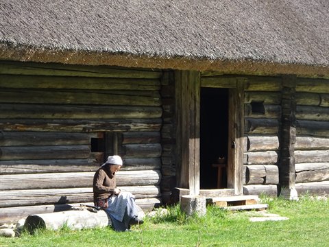 Woman in white headscarf sits outside log cabin with thatched roof