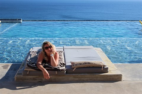 Woman in sunglasses lying on a daybed by the pool, blue sea behind