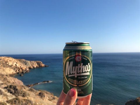 Mythos beer can raised in front of rocky Greek coastline
