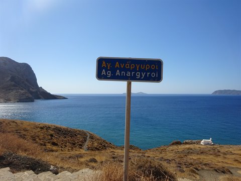 Blue road sign for Ag. Anargyroi above a turquoise bay with dry hills
