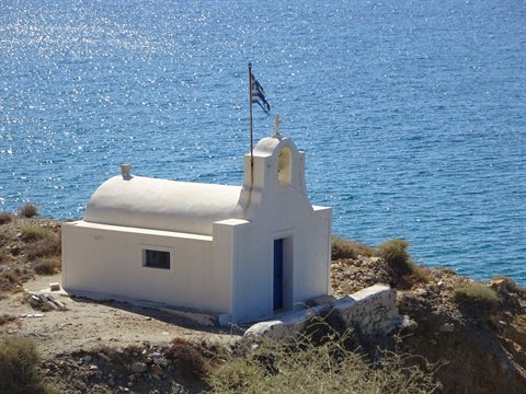 Whitewashed chapel with Greek flag on cliff above deep blue sea