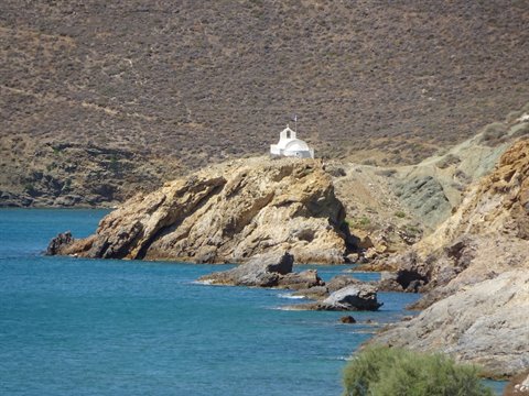 White chapel perched on rocky outcrop above turquoise sea, dry hills behind