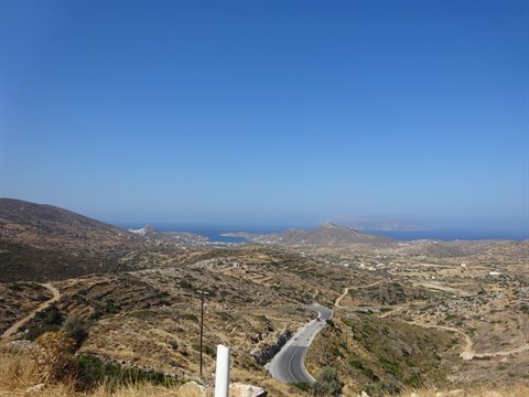 Winding road descending through dry hills toward white villages and blue sea