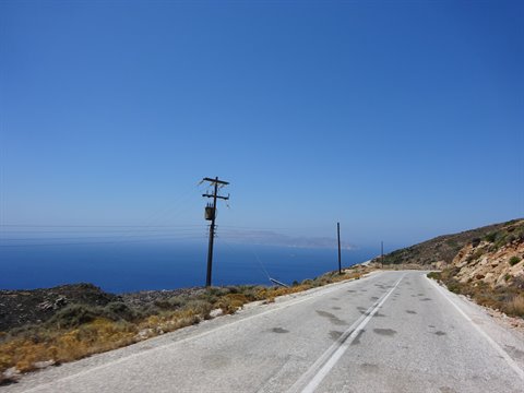 Asphalt road descends toward blue sea, wooden pole and dry shrubs under clear sky