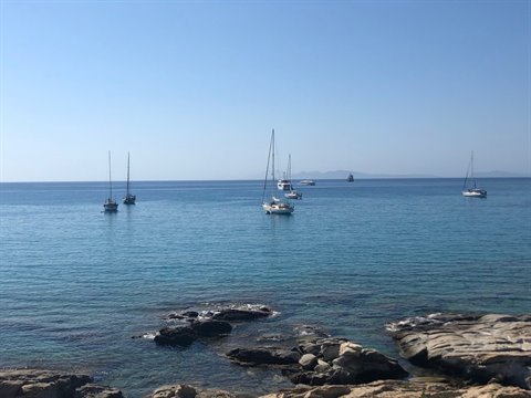 White sailboats anchored on calm turquoise sea, pale rocks in foreground, bright light