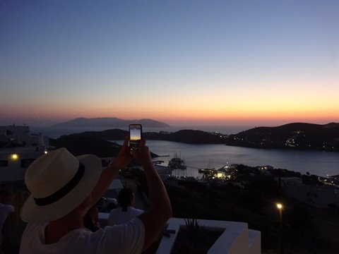 Tourist in hat photographs harbor at dusk with smartphone, lights glowing on water