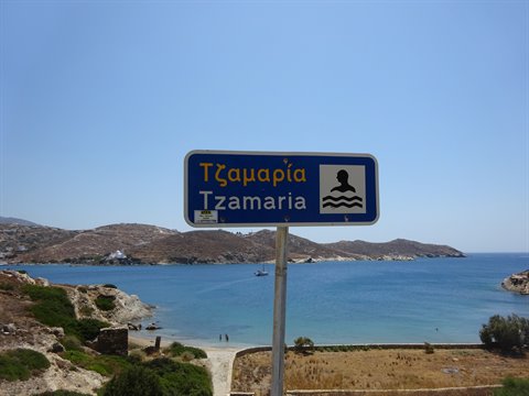 Blue road sign reading Tzamaria in front of a turquoise bay and dry hills