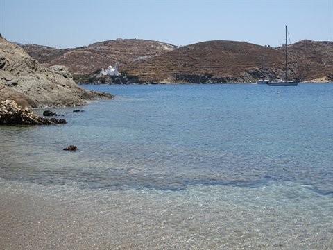 Bay with clear turquoise water, anchored sailboat, arid hills and white chapel, sunlight