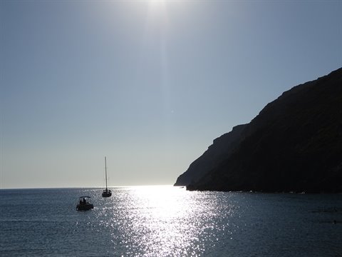 Boats anchored before the dark cliff, sun glittering on the Sifnos sea