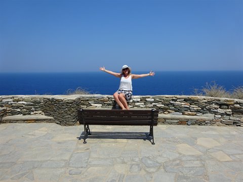 Tourist with open arms on a wooden bench by a stone wall overlooking the Aegean Sea