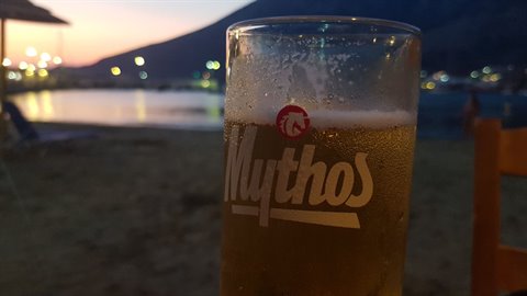 Glass of Mythos beer close-up, beach and harbor lights at dusk