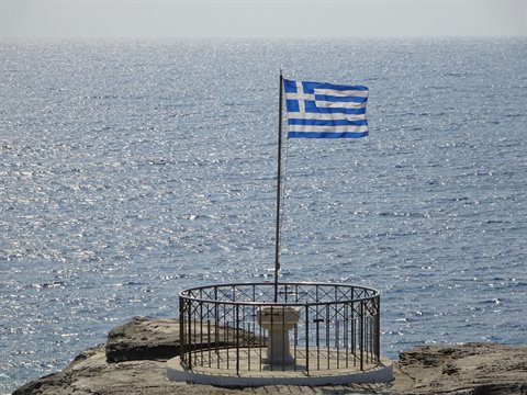 Greek flag waving over a circular iron platform on a Sifnos cliff
