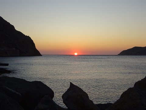 Red sun on the horizon between dark cliffs and rocks on a Sifnos cove
