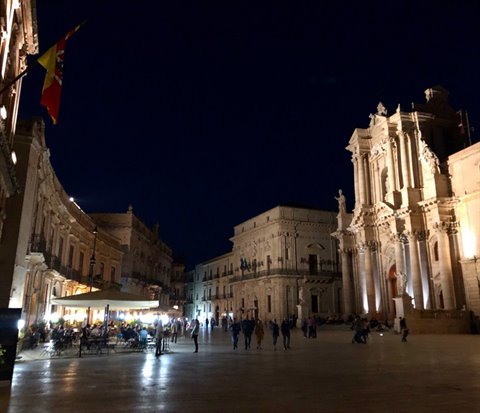 Piazza Duomo in Syracuse at night, people strolling among illuminated baroque buildings