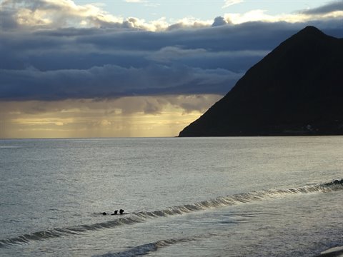 Swimmers on calm waves beneath a dark mountain and glowing clouds at Le Diamant